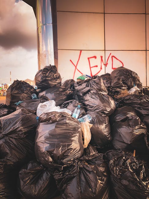 An alleyway between two buildings filled with a large amount of mixed waste, including several white and black trash bags, cardboard boxes, and loose debris scattered across the uneven, dark asphalt g