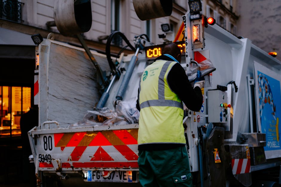 A sanitation worker wearing a high-visibility yellow vest and blue gloves is operating a white waste collection vehicle on an urban street during dusk. The vehicle's rear is open, revealing a load of